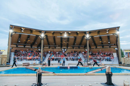 A lively outdoor arena with a large audience watching a lumberjack competition near a water feature.