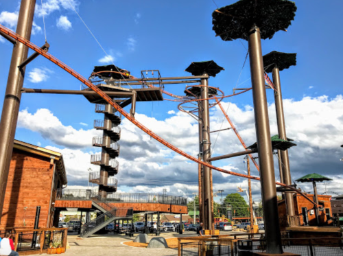 A large outdoor adventure park with tall structures, zip lines, and a spiral staircase under a blue sky with clouds.