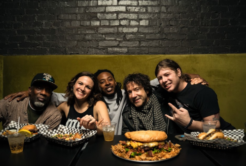 A group of five friends poses together at a restaurant, smiling with food and drinks on the table in front of them.
