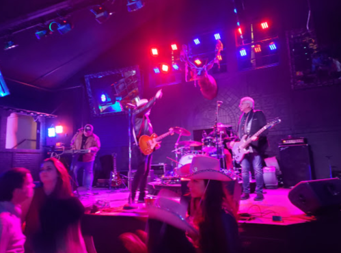 A lively band performs on stage with colorful lights, while people dance in the foreground wearing cowboy hats.