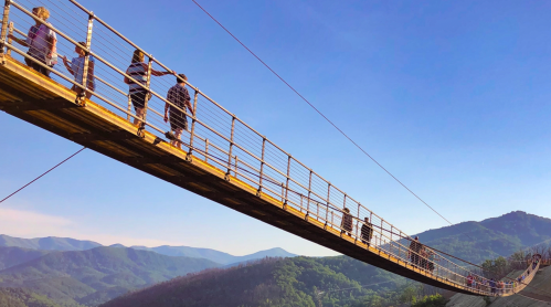 A group of people walking across a long wooden suspension bridge with mountains in the background under a clear blue sky.