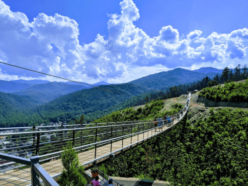A long suspension bridge stretches over a mountainous landscape under a blue sky with fluffy clouds.