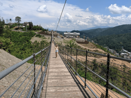 A person walks on a wooden suspension bridge with mountains and a blue sky in the background.