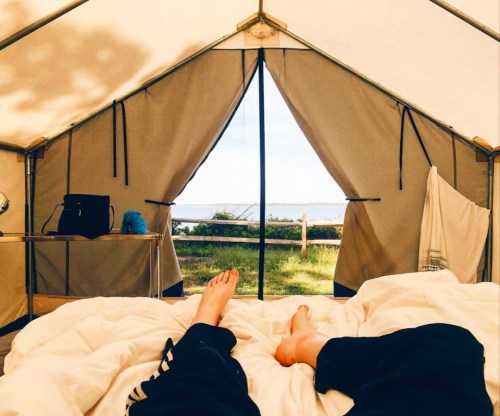 A view from inside a tent, showing feet on a bed with a scenic lake visible through the open entrance.