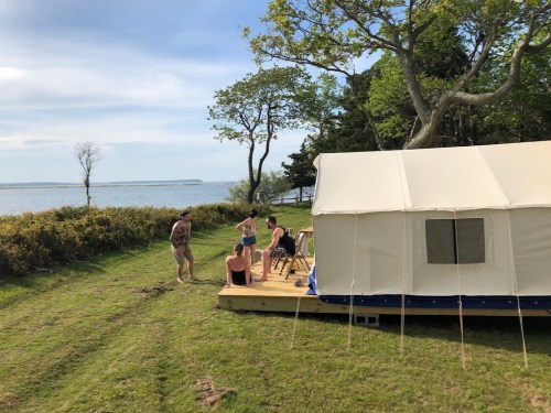 A group of people gather near a tent by the water, surrounded by greenery and trees on a sunny day.