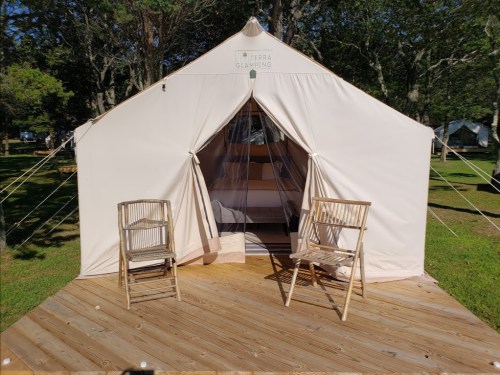 A beige glamping tent on a wooden deck, with two wooden chairs outside and trees in the background.