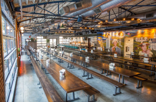 A spacious restaurant interior with long wooden tables, industrial decor, and a bar area in the background.