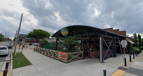 Exterior of a restaurant with a large awning, outdoor seating, and cloudy skies in the background.