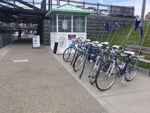 Bicycles lined up near a rental kiosk by a canal, with people walking and a bridge in the background.