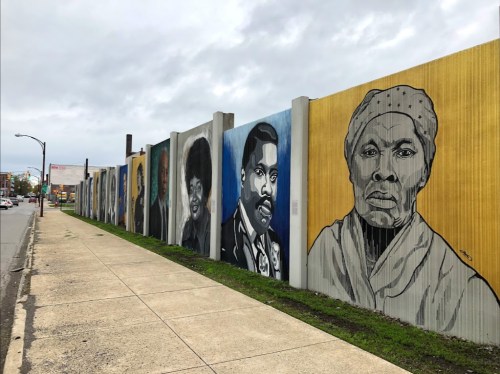 A colorful mural featuring portraits of notable figures on a concrete wall beside a sidewalk.