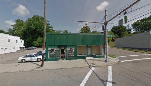 A green storefront at a street corner, with a traffic light and trees in the background on a sunny day.