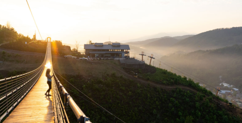 A person stands on a suspension bridge at sunset, with a scenic view of mountains and a building in the background.