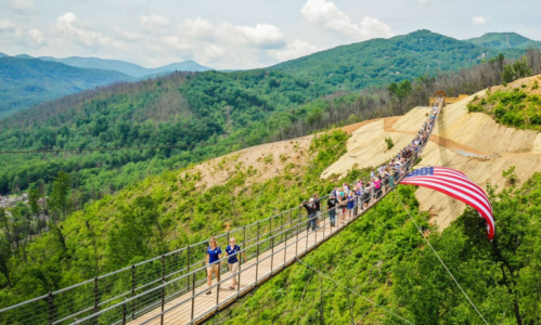 A long suspension bridge with people walking, surrounded by green mountains and a large American flag draping down.