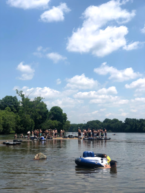 A sunny day at a river with people swimming and relaxing on inflatable floats, surrounded by trees and blue skies.