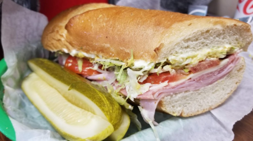 A sub sandwich filled with lettuce, tomato, and ham, served with a pickle on the side.