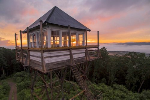 A wooden lookout tower with large windows, perched on stilts, against a vibrant sunset and misty landscape.