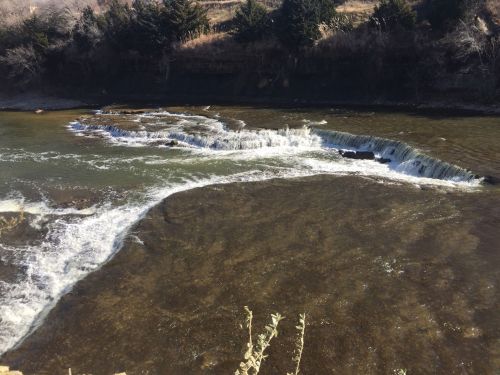 A serene river scene with gentle rapids and rocky banks, surrounded by trees and a hillside in the background.