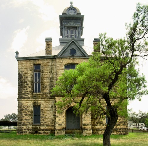 Historic stone building with a clock tower, surrounded by greenery and a tree, under a cloudy sky.