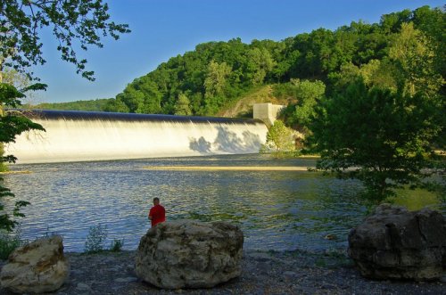 A person sits on rocks by a river, with a dam and lush green hills in the background under a clear blue sky.