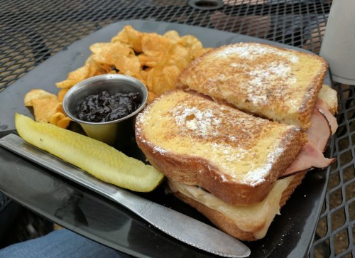 A plate with a sandwich, pickle, chips, and a small cup of jelly on a black table.