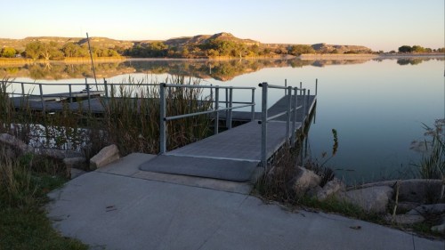 A calm lake at sunrise with a wooden dock and surrounding greenery, reflecting the serene landscape.