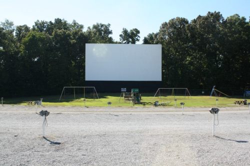 A drive-in movie theater with a large blank screen, swings, and picnic tables in a grassy area surrounded by trees.