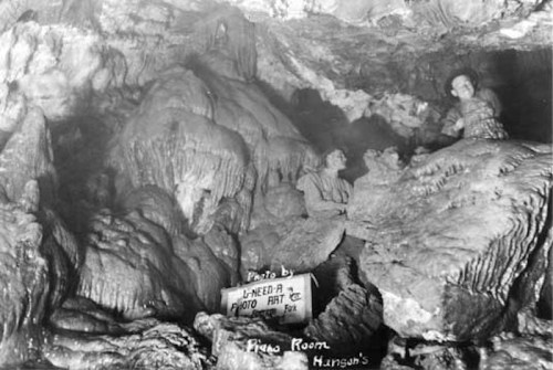 Black and white photo of a cave interior with stalactites and stalagmites, featuring two figures exploring the space.