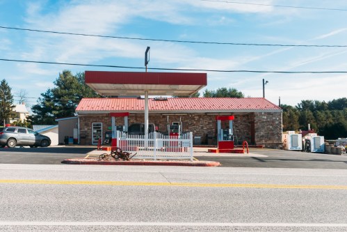 A small gas station with a red roof, stone facade, and white picket fence, set along a quiet road.