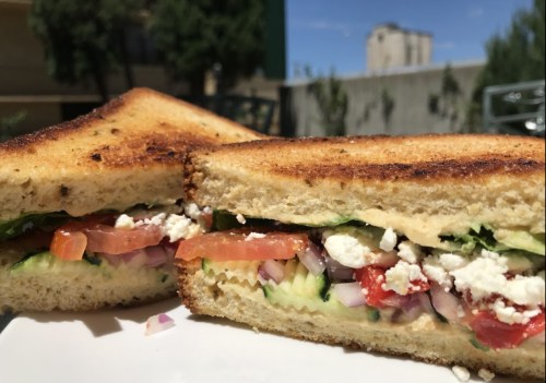A close-up of a toasted sandwich filled with vegetables and cheese, served on a white plate against a sunny background.