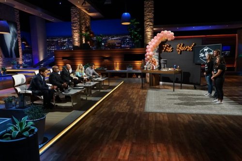A brightly lit studio with a panel of judges seated, two contestants standing, and a decorative backdrop featuring balloons.