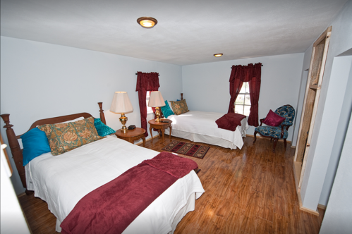Cozy bedroom with two beds, decorative pillows, lamps, and windows with red curtains. Warm wooden flooring.