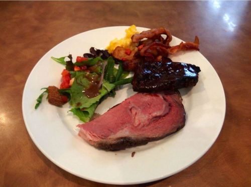 A plate with sliced roast beef, grilled meat, scrambled eggs, bacon, and a mixed green salad.