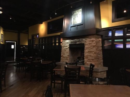 Cozy restaurant interior featuring wooden tables, black chairs, and a stone fireplace against a yellow wall.