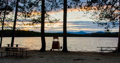 A serene lakeside scene at sunset, framed by trees, with picnic tables and a lifeguard chair in the foreground.