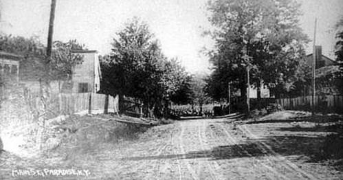 Black and white photo of a dirt road lined with trees and houses, capturing a quiet, rural scene.