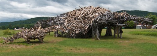 A large, abstract sculpture made of wooden planks, resembling a creature, set in a grassy field under a cloudy sky.