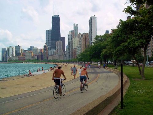 Two cyclists ride along a path by the lake, with a city skyline and beachgoers in the background.