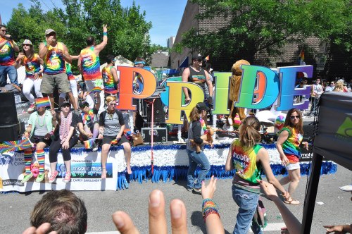 A vibrant parade float with colorful "PRIDE" letters, featuring people in rainbow attire celebrating LGBTQ+ pride.