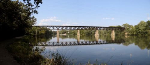 A railway bridge spans a calm river, surrounded by trees and a clear blue sky, with reflections on the water's surface.