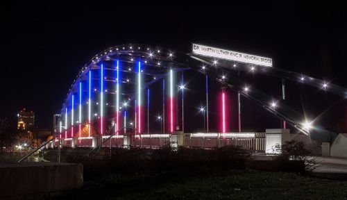 A brightly lit bridge at night, featuring red, white, and blue lights, with "Dr. Martin Luther King Jr. Memorial Bridge" sign.