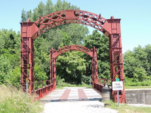 A red metal bridge with arched supports, surrounded by greenery, featuring a "Trail Closed" sign.