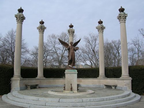 A bronze angel statue surrounded by tall columns in a circular stone plaza, set in a park with bare trees.