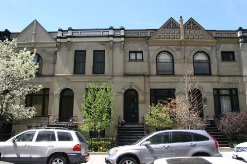 A row of historic townhouses with ornate architectural details, surrounded by greenery and parked cars.