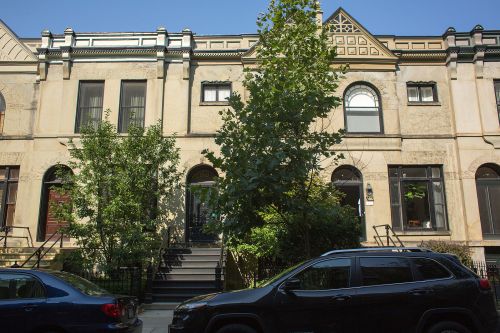 A row of historic townhouses with large windows and a tree in front, parked cars on the street below.