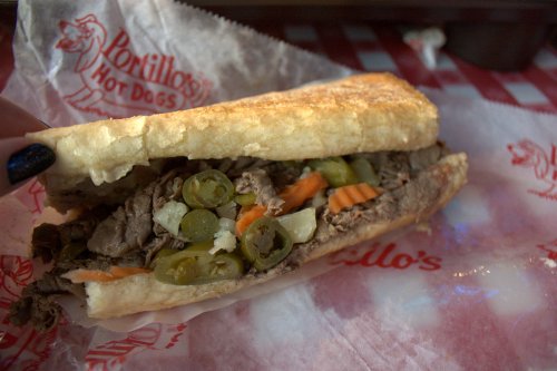 A close-up of a sandwich filled with beef, jalapeños, and vegetables, served in a paper wrapper.