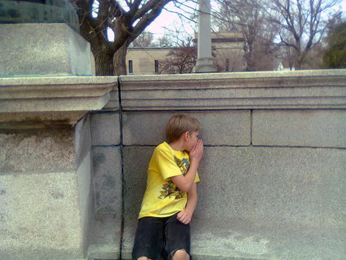 A young boy in a yellow shirt sits against a stone wall, whispering while looking away. Trees and a lamppost are in the background.