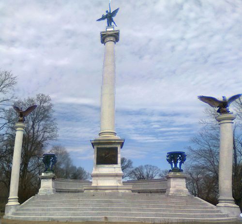 A tall monument with a central column, topped by a statue, flanked by eagles and decorative urns, set against a cloudy sky.