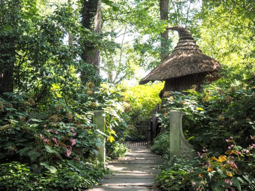 A stone pathway leads through lush greenery to a whimsical thatched-roof structure in a serene garden.