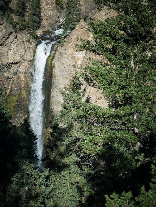 A waterfall cascades down rocky cliffs, surrounded by lush green trees in a natural landscape.