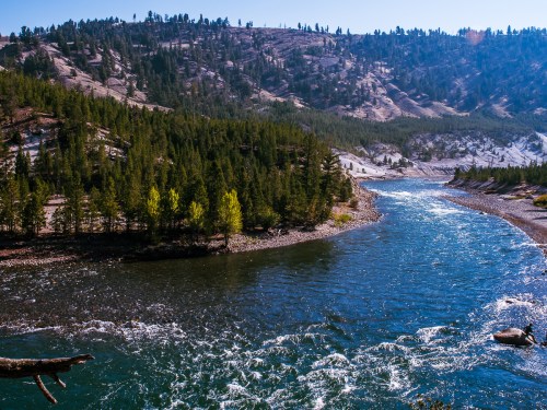 A winding river flows through a forested landscape, surrounded by mountains under a clear blue sky.
